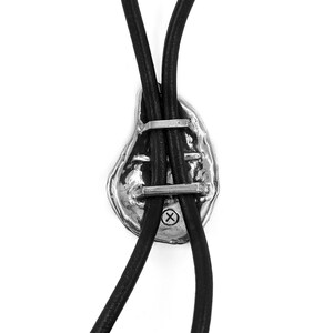 May include: A silver bolo tie with a textured, abstract design. The tie is attached to two black leather cords.