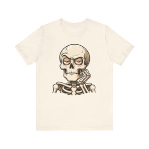 Może przedstawiać: Kremowy t-shirt z krótkim rękawem z grafiką przedstawiającą szkielet z kreskówki o pomarańczowych oczach. Szkielet ma zamyślony wyraz twarzy, opiera podbródek na dłoni i jest utrzymany w odcieniach beżu oraz brązu.