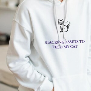 May include: A white hooded sweatshirt featuring a black line drawing of a cat and the purple text "STACKING ASSETS TO FEED MY CAT" printed on the chest. The garment includes a front kangaroo pocket, long sleeves, and a drawstring hood.