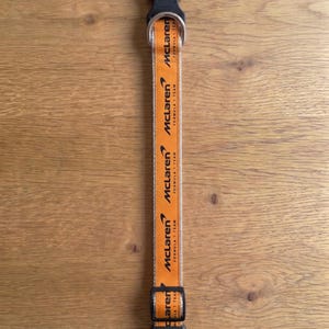 May include: An orange pet collar featuring the black McLaren Formula 1 Team logo and text repeated along the length of the strap. The collar includes a black plastic side-release buckle, a metal D-ring for leash attachment, and a black plastic slider.