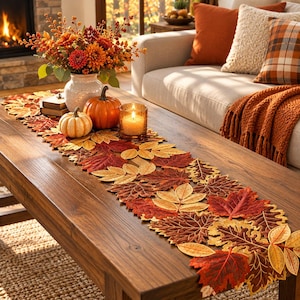 May include: A long table runner featuring a dense arrangement of fabric autumn leaves in shades of red, orange, yellow, and brown. The runner is laid flat on a wooden table, decorated with a white vase of fall flowers, two pumpkins, and a candle.