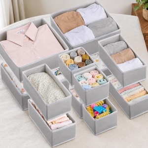 May include: A set of light gray fabric storage organizers in various rectangular and square sizes. The containers hold folded clothing items including shirts, socks, and underwear, as well as a small collection of colorful plastic building blocks.