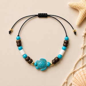 May include: A bracelet featuring a central turquoise-coloured turtle charm, flanked by dark brown wooden beads, bright turquoise round beads, white cylindrical beads, and small gold-toned metal accents on a black adjustable cord with gold-toned end beads.