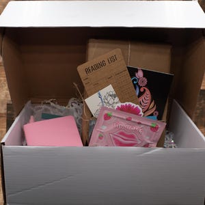 May include: A white cardboard box containing a pink notebook, a brown paper-wrapped package, a tan card labeled READING LIST, a white card with a floral line drawing, a pink strawberry-themed lip mask packet, and a black card with floral art.