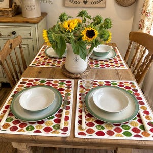 May include: A wooden dining table set for two with placemats featuring a colourful autumn leaf pattern. Each place setting includes a white bowl, a green-rimmed plate, a fork, and a knife. A white pitcher filled with sunflowers sits in the centre.