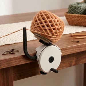 Op de afbeelding: Een garenwinder met een witte ronde behuizing, een zwarte hendel en een zwarte verticale geleidearm. Het apparaat is aan de rand van een tafel geklemd en houdt een grote, beige bol garen vast die door het geleidemechanisme wordt opgewonden.