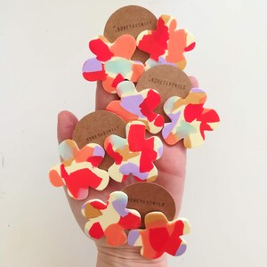 May include: Five colorful flower-shaped earrings with a white background and a red, orange, yellow, purple, and blue abstract pattern. The earrings are made by Honeyandmilk.