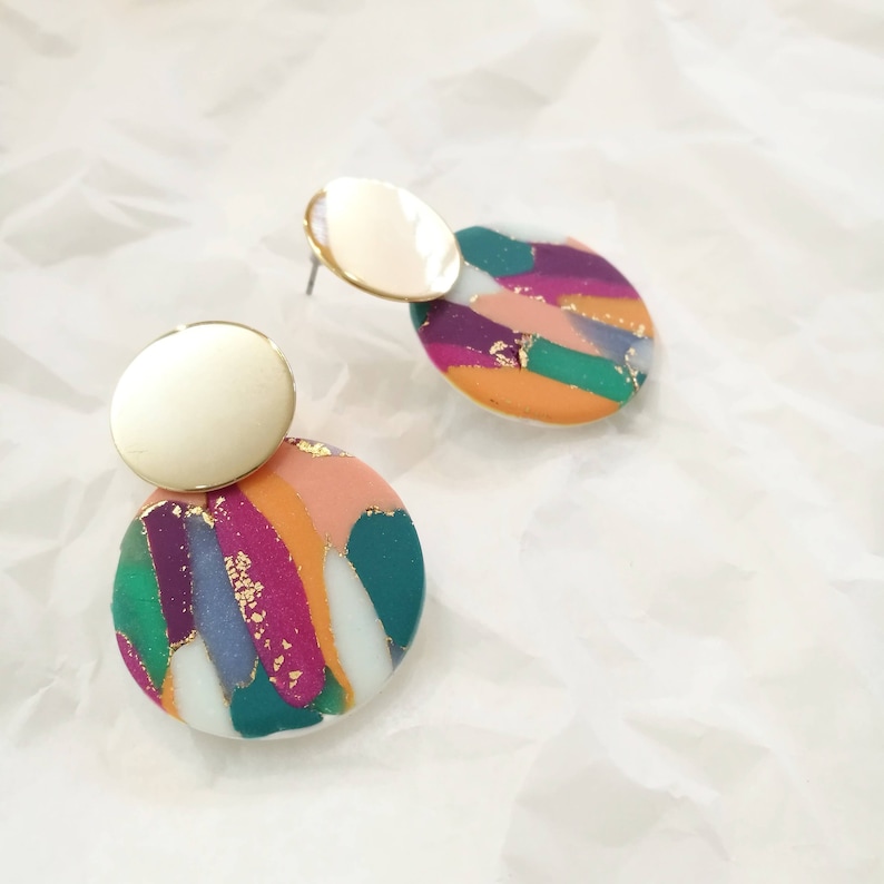 May include: A pair of gold and colorful abstract patterned earrings. The earrings are round and have a gold disc on top of the patterned disc.