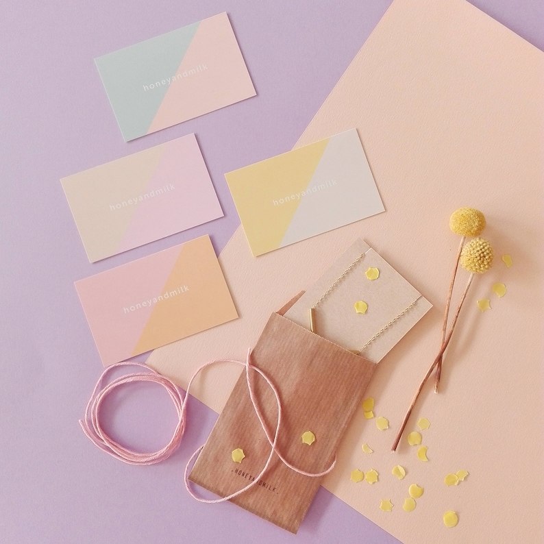 May include: A brown paper envelope with the text "honeyandmilk" on it. The envelope is surrounded by small yellow confetti and a few yellow flowers. There are four pink and white business cards with the text "honeyandmilk" on them.
