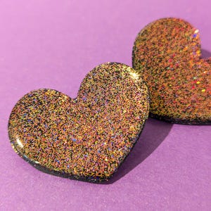 Op de afbeelding: Twee zwarte hartvormige harsversieringen met gouden en iriserende glitter ingebed in de hars.