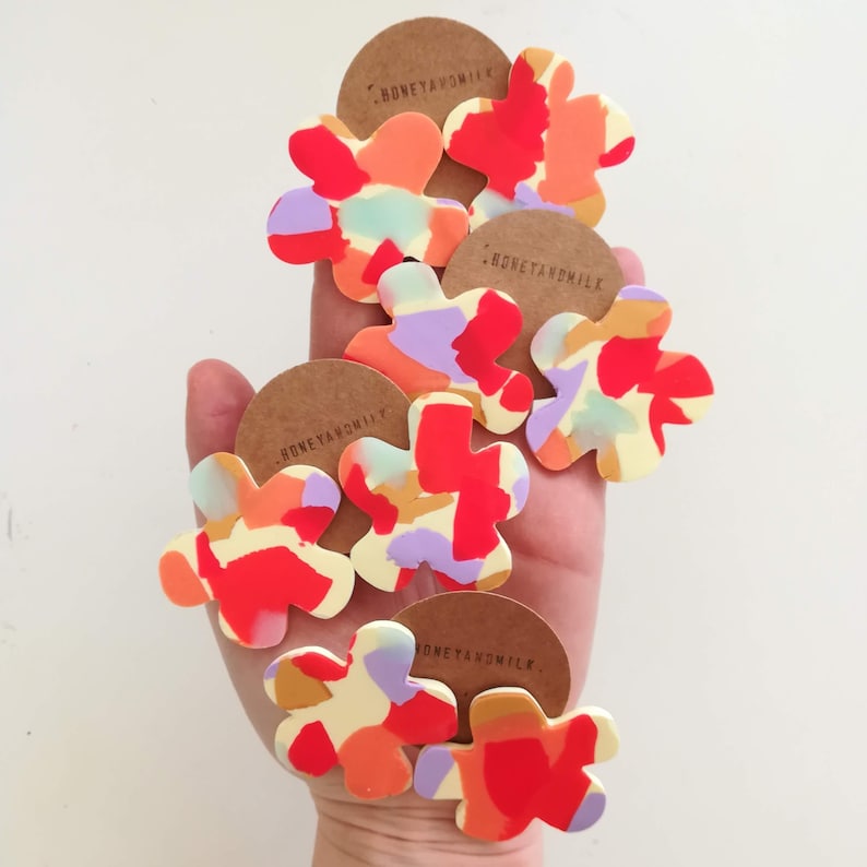 May include: Five colorful flower-shaped earrings with a white background and a red, orange, yellow, purple, and blue abstract pattern. The earrings are made by Honeyandmilk.