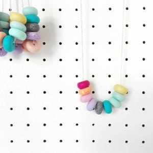May include: A rainbow coloured necklace with round beads made of polymer clay. The necklace is on a silver chain and is hanging on a white pegboard.