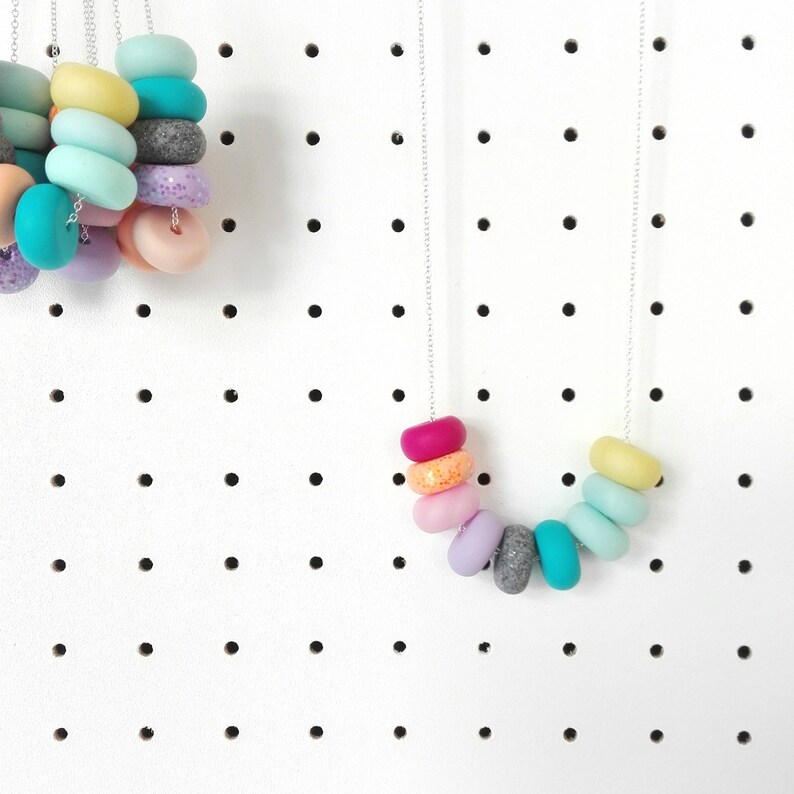 May include: A rainbow coloured necklace with round beads made of polymer clay. The necklace is on a silver chain and is hanging on a white pegboard.