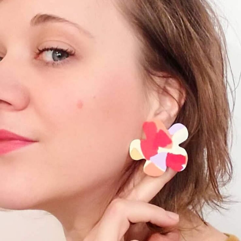 May include: A pair of colorful abstract flower-shaped earrings. The earrings are made of polymer clay and have a white, pink, orange, and purple color scheme.