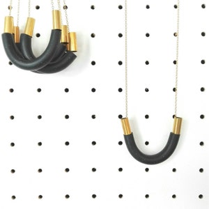 Op de afbeelding: Twee kettingen met zwarte rubberen en gouden metalen accenten. De kettingen worden getoond op een wit prikbord.