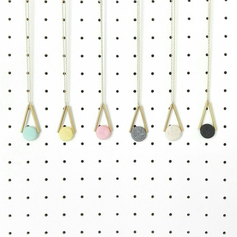 Op de afbeelding: Zes gouden kettingen met verschillende ronde hangers in verschillende kleuren die aan een wit prikbord hangen. De hangers zijn lichtblauw, geel, roze, grijs, wit en zwart.