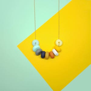 Op de afbeelding: Een kleurrijke ketting met een gouden ketting en een verscheidenheid aan kralen in tinten blauw, grijs, geel, oranje en wit. De kralen zijn in een gebogen vorm gerangschikt, waardoor een uniek en opvallend ontwerp ontstaat. De ketting wordt gepresenteerd tegen een gele en teal achtergrond.