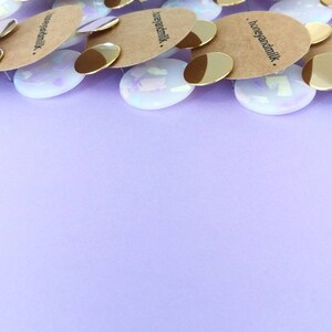 May include: A close-up of several pairs of iridescent white earrings with gold accents. The earrings are stacked on top of each other and have the text "honeyandmilk." printed on the back.