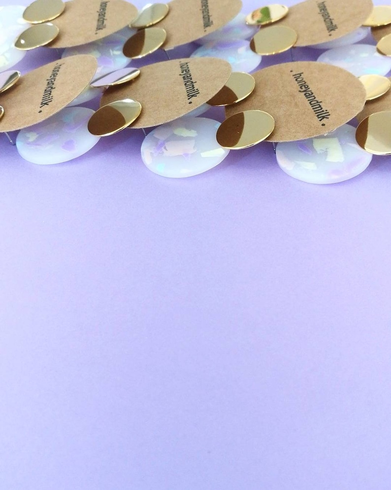 May include: A close-up of several pairs of iridescent white earrings with gold accents. The earrings are stacked on top of each other and have the text "honeyandmilk." printed on the back.