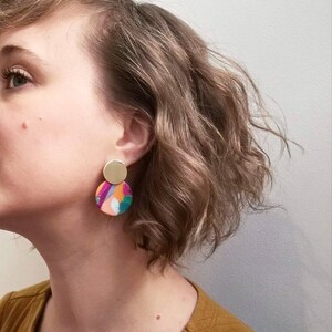 May include: A pair of colorful, abstract earrings with a gold accent. The earrings are round and feature a variety of colors, including pink, orange, green, and purple.