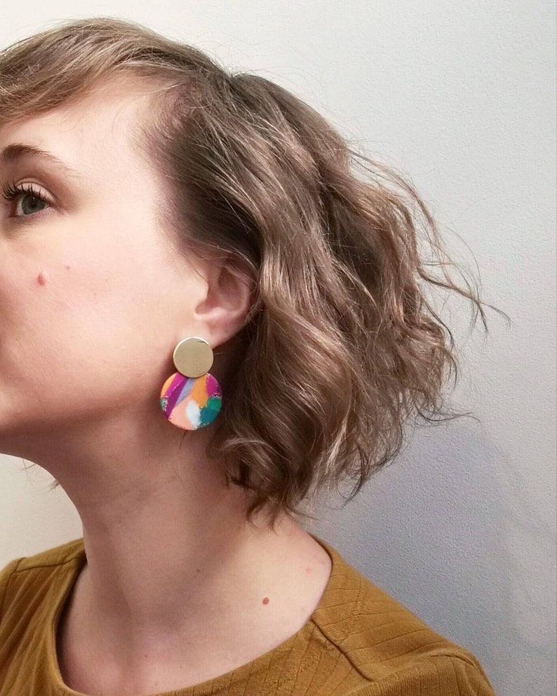 May include: A pair of colorful, abstract earrings with a gold accent. The earrings are round and feature a variety of colors, including pink, orange, green, and purple.