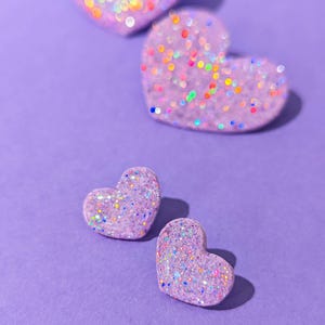 Op de afbeelding: Verschillende glitterende, lavendelkleurige hartvormige decoraties zijn verspreid op een paarse achtergrond. De harten zijn bedekt met iriserende glitter, die verschillende kleuren reflecteert. De afbeelding is een close-up, met harten van verschillende grootte.