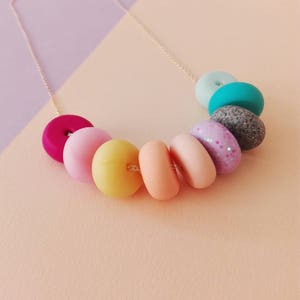 May include: A colourful necklace with pastel-coloured donut-shaped beads. The beads are in shades of pink, yellow, orange, turquoise, and grey. The necklace is made with a gold chain.