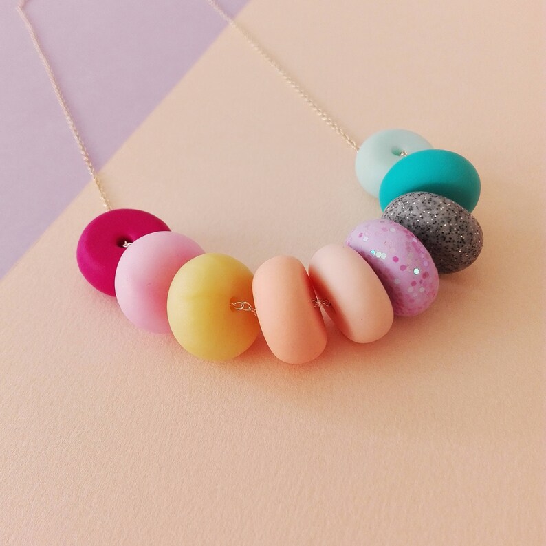 May include: A colourful necklace with pastel-coloured donut-shaped beads. The beads are in shades of pink, yellow, orange, turquoise, and grey. The necklace is made with a gold chain.