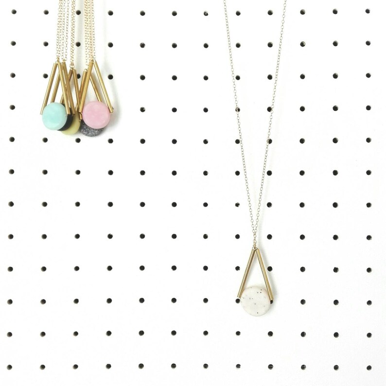 Op de afbeelding: Drie gouden kettingen met verschillende ronde hangers in verschillende kleuren hangen aan een wit prikbord. De hangers zijn lichtblauw, roze en grijs.