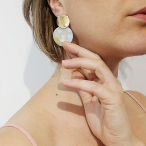 May include: A pair of gold and white statement earrings with a round shape. The earrings have a gold disc on top of a larger white disc with a subtle iridescent finish.