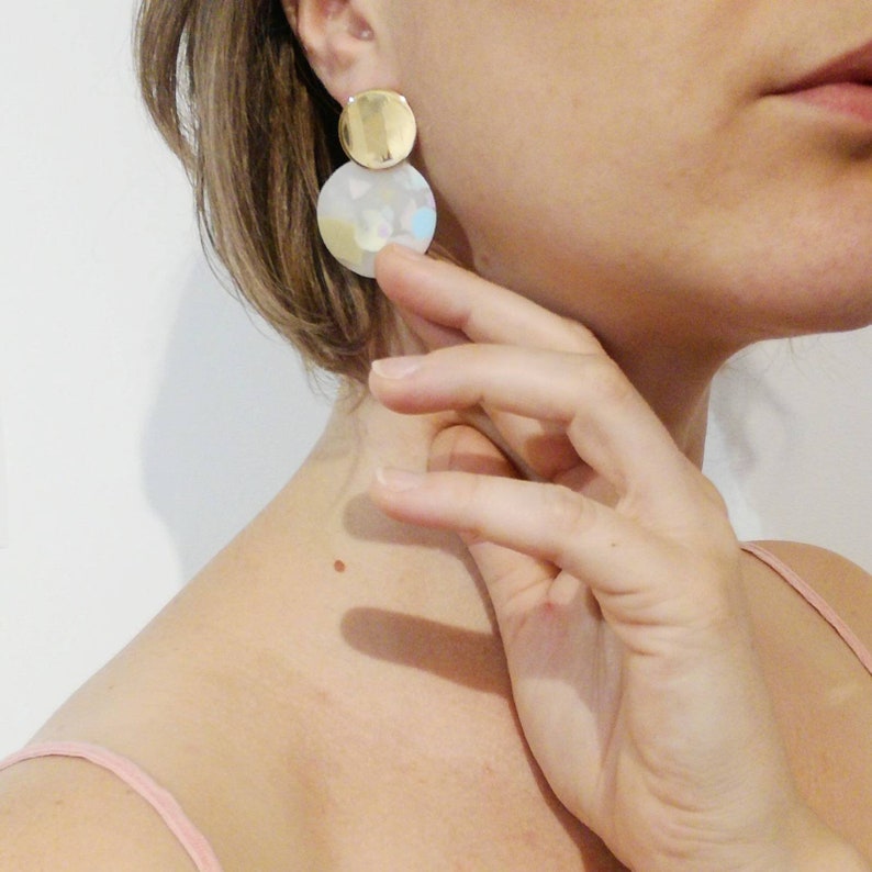 May include: A pair of gold and white statement earrings with a round shape. The earrings have a gold disc on top of a larger white disc with a subtle iridescent finish.