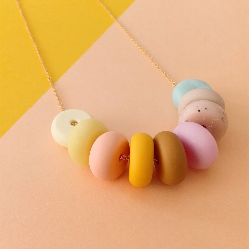 May include: A gold chain necklace with pastel-coloured donut-shaped beads. The beads are in a rainbow order, starting with white, then yellow, peach, orange, brown, pink, light pink, and light blue.