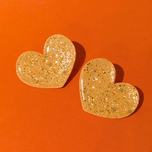 Op de afbeelding: Twee hartvormige objecten met een glinsterend, goudgeel oppervlak tegen een oranje achtergrond. De harten hebben een glanzende afwerking en zijn bedekt met gouden glitter. De afbeelding is goed verlicht en benadrukt de textuur en glans.