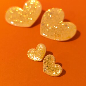 Op de afbeelding: Vier gouden glitterhartvormige ornamenten op een oranje achtergrond. De harten zijn van verschillende grootte en zijn in een cluster gerangschikt.