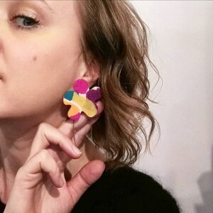May include: A pair of colorful flower-shaped earrings. The earrings are made of a variety of materials, including paper, fabric, and paint. The earrings are a bright pink, yellow, orange, green, and purple.