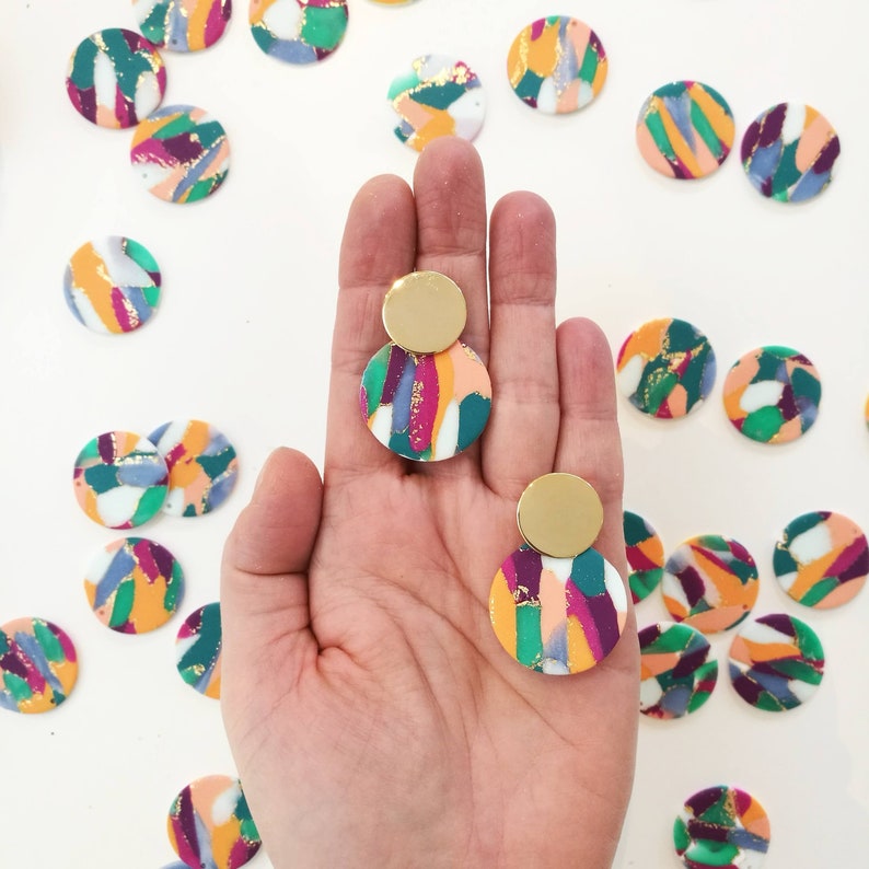 May include: A pair of colorful, abstract patterned earrings with gold accents. The earrings are made of polymer clay and feature a round shape with a mix of pink, green, white, and gold colors.