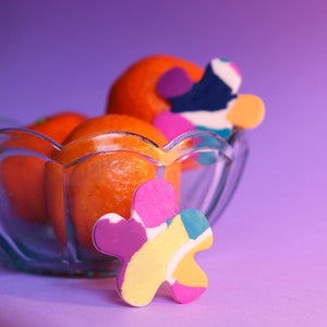 May include: A glass bowl filled with orange mandarins, with a colorful abstract flower-shaped object in the foreground. The bowl and fruit are on a purple background.