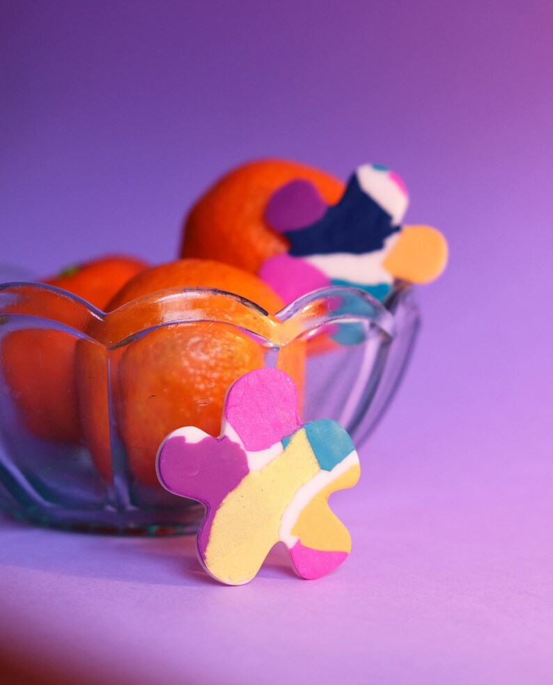 May include: A glass bowl filled with orange mandarins, with a colorful abstract flower-shaped object in the foreground. The bowl and fruit are on a purple background.
