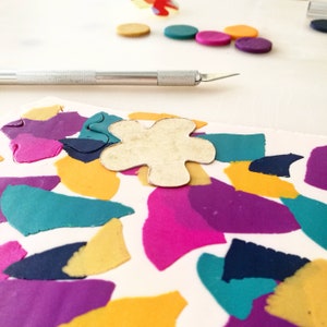May include: A close-up of a craft project with a flower-shaped cutout on top of a sheet of colorful polymer clay. The clay is arranged in a mosaic pattern with shades of purple, pink, teal, yellow, and navy blue.