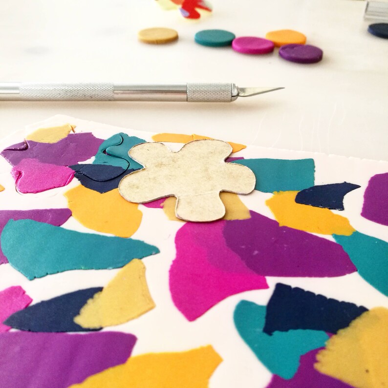 May include: A close-up of a craft project with a flower-shaped cutout on top of a sheet of colorful polymer clay. The clay is arranged in a mosaic pattern with shades of purple, pink, teal, yellow, and navy blue.