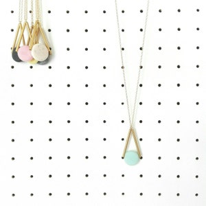 Op de afbeelding: Een gouden geometrische ketting met een lichtblauwe ronde hanger. De ketting hangt aan een prikbord met andere kettingen.