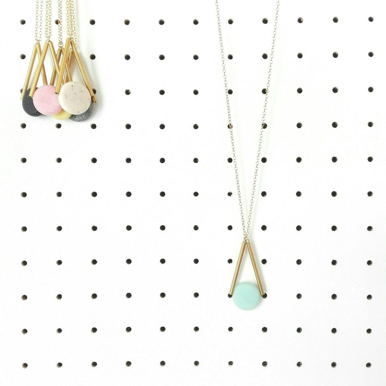 Op de afbeelding: Een gouden geometrische ketting met een lichtblauwe ronde hanger. De ketting hangt aan een prikbord met andere kettingen.