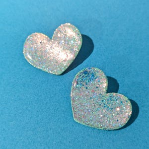 Op de afbeelding: Twee glinsterende hartvormige harsversieringen met iriserende glitter. De harten zijn lichtblauw en wit.