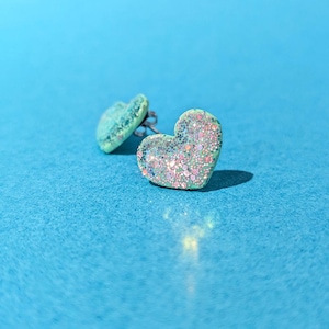 Op de afbeelding: Een paar hartvormige oorstekers. Eén oorbel is scherp, met een lichtgroene basis bedekt met iriserende glitter. De andere oorbel is iets onscherp. De oorbellen staan tegen een helderblauwe achtergrond.