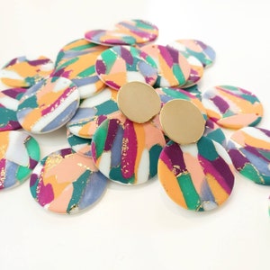 May include: A pile of colorful round polymer clay earrings with gold accents. The earrings are painted with abstract patterns in shades of pink, purple, green, blue, and gold. Two gold earring posts are visible in the center of the image.