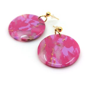 Op de afbeelding: Een paar ronde oorbellen met een marmer effect in roze en paars, met gouden accenten. De oorbellen hebben goudkleurige oorhaken en -stiften. De oorbellen staan tegen een witte achtergrond.