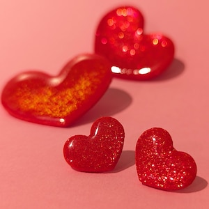 Op de afbeelding: Vier rode hartvormige ornamenten met glitter. Twee van de harten zijn kleiner en twee zijn groter. De grotere harten hebben een meer oranje tint.