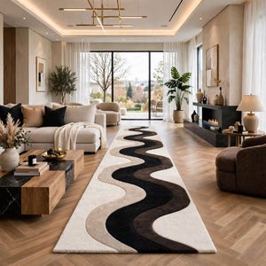 Könnte beinhalten: Ein langer rechteckiger Teppich mit einem sich wiederholenden Wellenmuster in Creme-, Beige- und Schwarztönen. Der Teppich liegt auf einem hellbraunen Fischgrät-Holzboden in einem Wohnzimmer mit Sofa, Couchtisch und Sessel.
