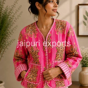May include: A bright pink quilted jacket featuring a repeating tiger pattern and vertical white stripes. The jacket has a V-neckline, long sleeves with folded cuffs, and two front patch pockets. The text jaipuri exports is printed in white.