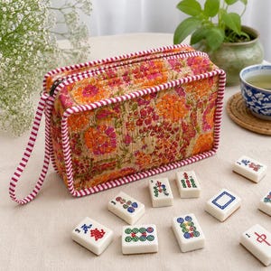 Könnte beinhalten: Eine rechteckige, gesteppte Stofftasche mit einem leuchtend orange-rosa Blumenmuster, rotem und weißem Streifenbesatz und einer passenden Handschlaufe, begleitet von mehreren weißen Mahjong-Spielsteinen mit bunten Gravuren.